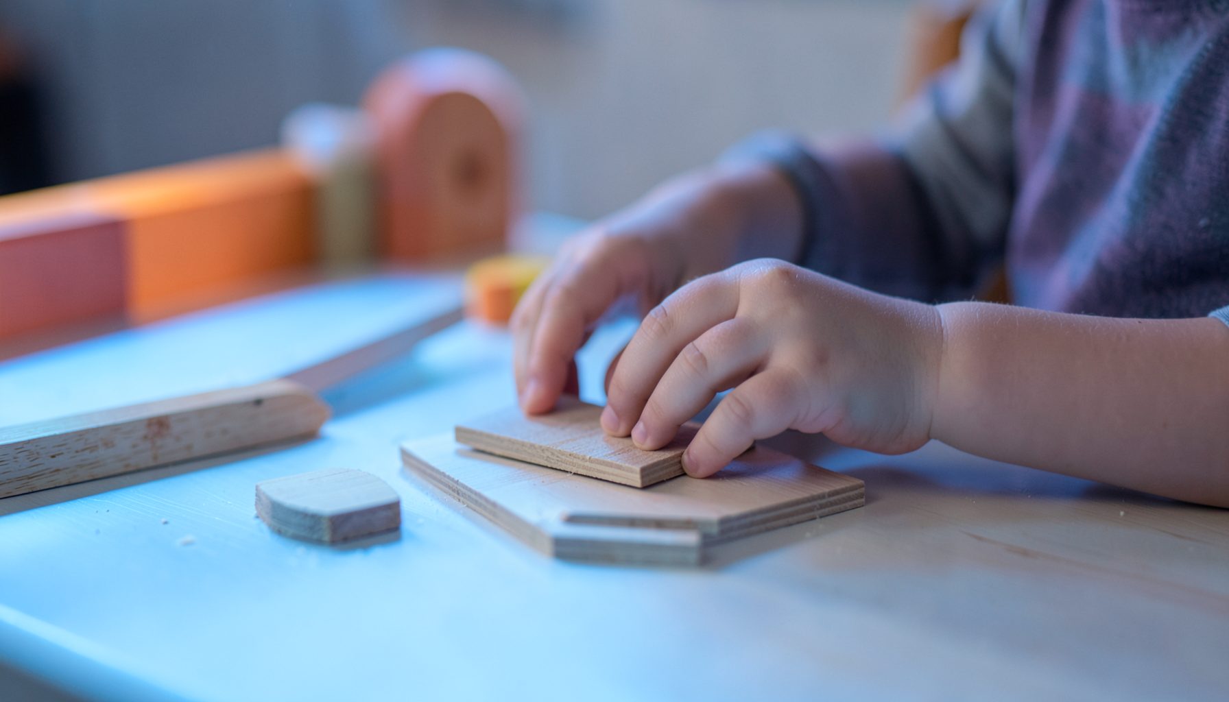 Manos infantiles manipulando piezas de madera sobre una mesa de luz con tonos azulados y anaranjados suaves, en un entorno de juego tranquilo
