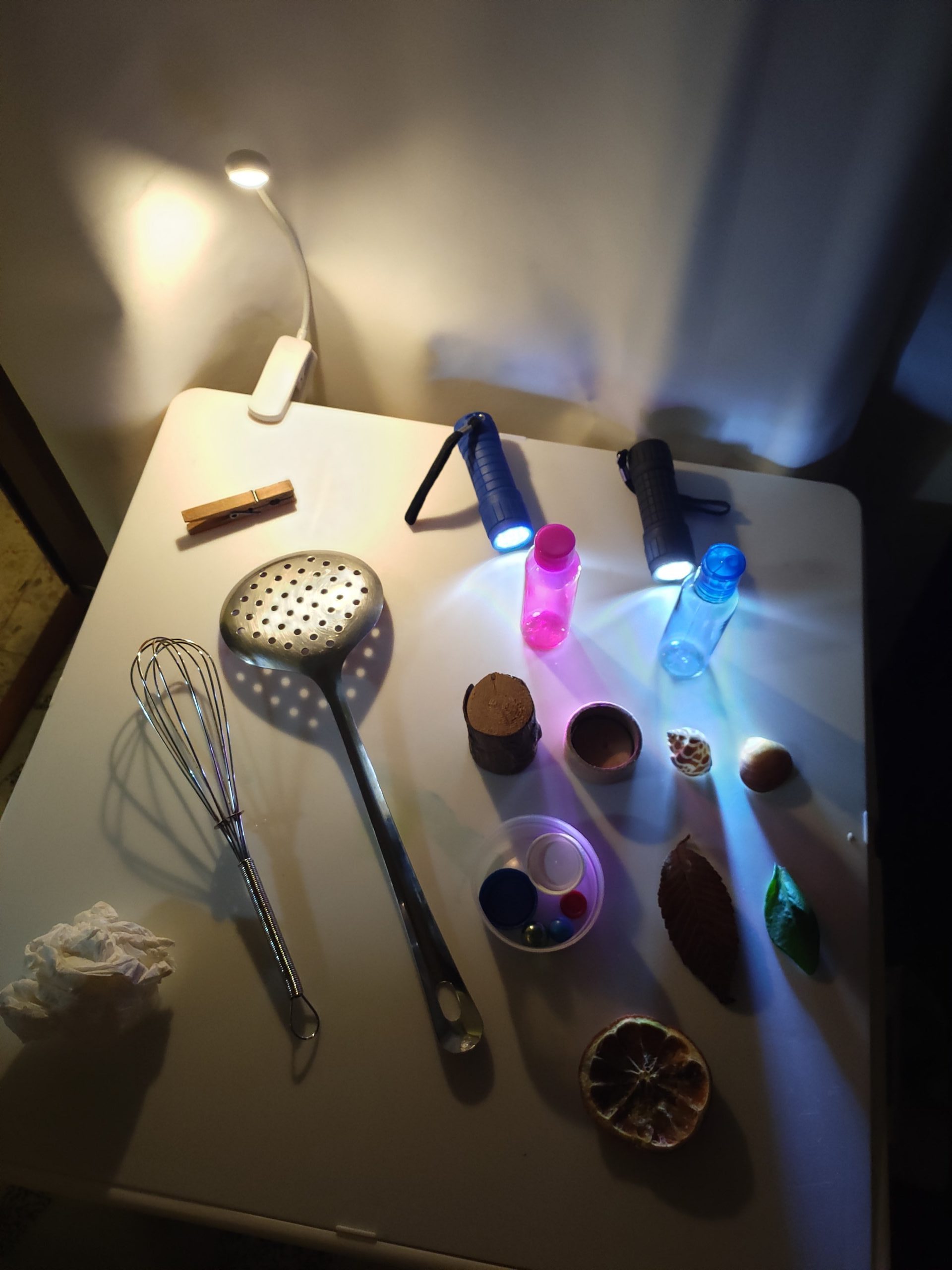Vista cenital de una mesa con herramientas de cocina, pequeños frascos, linternas, conchas y una hoja, iluminada por haces de luz de colores.